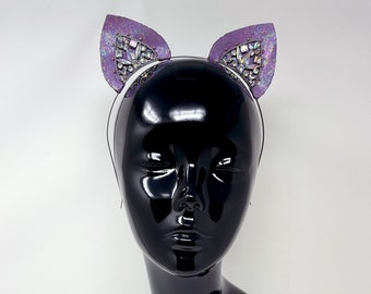 May include: A black mannequin head wearing a purple and iridescent cat ear headband. The headband is made of fabric and has a white elastic band.