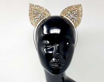 May include: A white headband with gold cat ears adorned with rhinestones. The headband is on a black mannequin head.