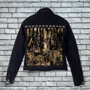 May include: Black denim jacket with a gold sequin and leopard print back panel. The back panel features fringe and is accented with silver eyelets. The jacket has a classic collar, button closures, and long sleeves.