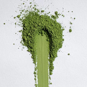 May include: A close-up shot of vibrant green matcha powder on a white surface. A streak of matcha powder is dragged down the center, with scattered granules around it. The image highlights the texture and color of the finely ground tea.