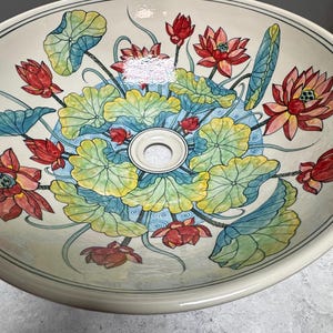 Puede incluir: Un lavabo de cerámica blanca con un diseño floral que presenta flores de loto rosas y rojas y hojas verdes. El lavabo tiene forma redonda y un agujero de drenaje en el centro.