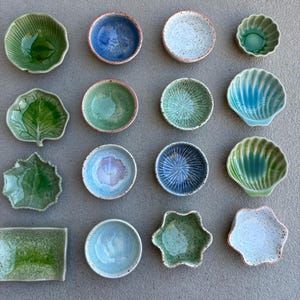 May include: A collection of small, handcrafted ceramic dishes in various shapes and colors. The dishes include leaf-shaped, round, and shell-shaped designs, with shades of green, blue, and white. Some have textured surfaces and speckled finishes, perfect for serving small portions.