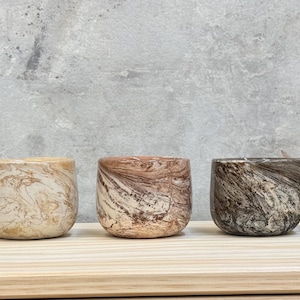 May include: Five small, round, handleless cups arranged in a row. Each cup features a distinct marbled pattern with swirling colors: blue and white, beige and cream, reddish-brown and cream, dark brown and cream, and sage green and cream.