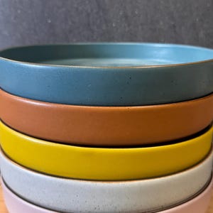 May include: A stack of five ceramic bowls in various colors, including teal, orange, yellow, light gray, and pink. The bowls are stacked on a wooden surface.