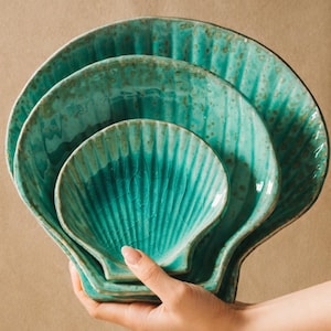 May include: A set of three turquoise seashell-shaped ceramic dishes. The dishes are nested together, with the largest in the back and the smallest in the front. The dishes have a textured, ridged surface and a distressed finish. The text "CHEF'S TABLEWARE" is at the bottom.
