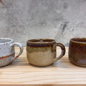 May include: Three ceramic mugs with handles. The left mug is white with brown speckles and a reddish-brown band. The middle mug is tan with brown speckles and a dark brown rim. The right mug is a solid dark brown with a glossy, dripped glaze finish.