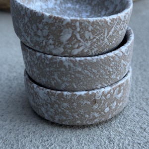 May include: A stack of three small, round ceramic bowls. The bowls have a speckled, textured surface with a light brown base and white accents. The bowls are stacked on top of each other.