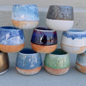 May include: A collection of handmade ceramic stemless wine glasses. Each glass features a unique color scheme, including blue, green, and gold, with speckled orange bases. The glasses are stacked in a visually appealing arrangement.