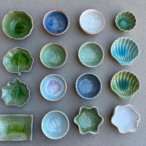 May include: An assortment of small, handcrafted ceramic bowls in various shapes and colors. The bowls include leaf, shell, and floral designs, with shades of green, blue, and white. Some bowls have textured surfaces and unique glaze patterns.