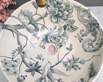 Könnte beinhalten: Rundes Waschbecken aus cremefarbener Keramik mit floralen und Vogelmotiven in Blau- und Grautönen. Das Waschbecken hat ein zentrales Abflussloch mit rosa Akzent und wird durch eine goldfarbene Armatur ergänzt.