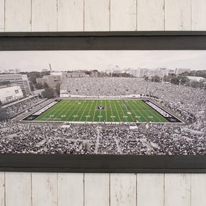 May include: Framed black and white photograph of a football stadium filled with spectators. The field is green with white yard lines and end zones. The frame is a dark gray color.