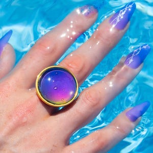 Large gold circle mood ring worn on hand in water from swimming pool, long purple nails on hand