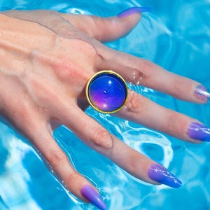 Large gold circle mood ring worn on hand in water from swimming pool, long purple nails on hand
