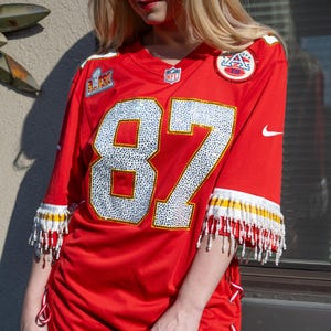 Puede incluir: Camiseta de f&uacute;tbol roja con el n&uacute;mero 87 en blanco y plata. La camiseta tiene ribetes blancos y amarillos con flecos de cuentas en las mangas. La camiseta tiene detalles de cinta roja y blanca en los laterales.