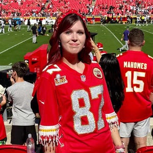 Puede incluir: Un vestido de camiseta de f&uacute;tbol roja con el n&uacute;mero 87 en plata. El vestido tiene flecos blancos y dorados en las mangas. El fondo muestra un estadio de f&uacute;tbol con aficionados y jugadores. El vestido se lleva con sandalias.