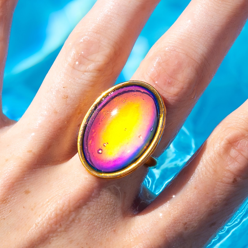 gold waterproof mood ring large oval shape, purple, pink, yellow mood stone, worn on hand swimming in water from a swimming pool. Cropped image for google shopping search.