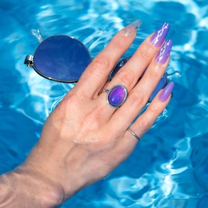 A silver ring with a small, oval, purple blue color changing stone. The ring is on a hand with long, purple fingernails holding sunglasses, set against a backdrop of blue water. The water's surface reflects light, creating a shimmering effect.