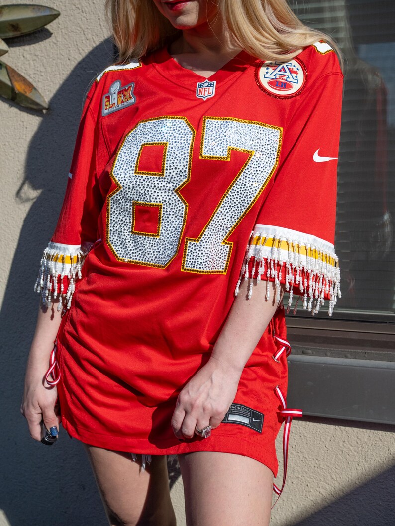 Puede incluir: Camiseta de f&uacute;tbol roja con el n&uacute;mero 87 en blanco y plata. La camiseta tiene ribetes blancos y amarillos con flecos de cuentas en las mangas. La camiseta tiene detalles de cinta roja y blanca en los laterales.