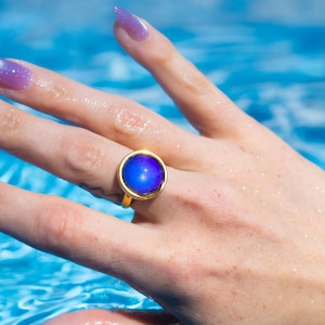 Small gold circle mood ring worn on hand in water from swimming pool, long purple nails on hand