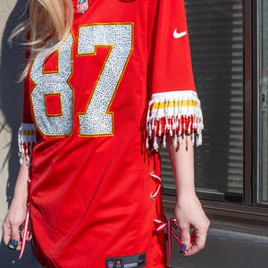 Puede incluir: Camiseta de f&uacute;tbol roja con el n&uacute;mero 87 en blanco, adornada con pedrer&iacute;a. La camiseta tiene flecos blancos y amarillos en las mangas y cordones de cinta rojos y blancos en los laterales. Con logotipos de la NFL y Nike.