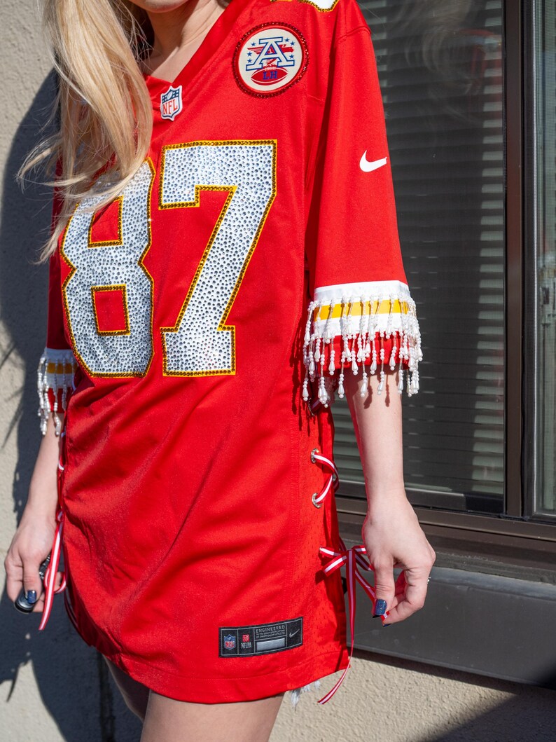 Puede incluir: Camiseta de f&uacute;tbol roja con el n&uacute;mero 87 en blanco, adornada con pedrer&iacute;a. La camiseta tiene flecos blancos y amarillos en las mangas y cordones de cinta rojos y blancos en los laterales. Con logotipos de la NFL y Nike.