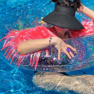 Waterproof mood ring and waterproof mood bracelet worn on model wearing a neon pink tinsel fringe karma jacket floating in a pool on a glitter filled transparent donut shaped raft