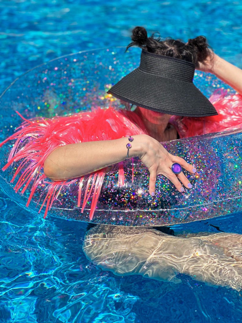 Waterproof mood ring and waterproof mood bracelet worn on model wearing a neon pink tinsel fringe karma jacket floating in a pool on a glitter filled transparent donut shaped raft