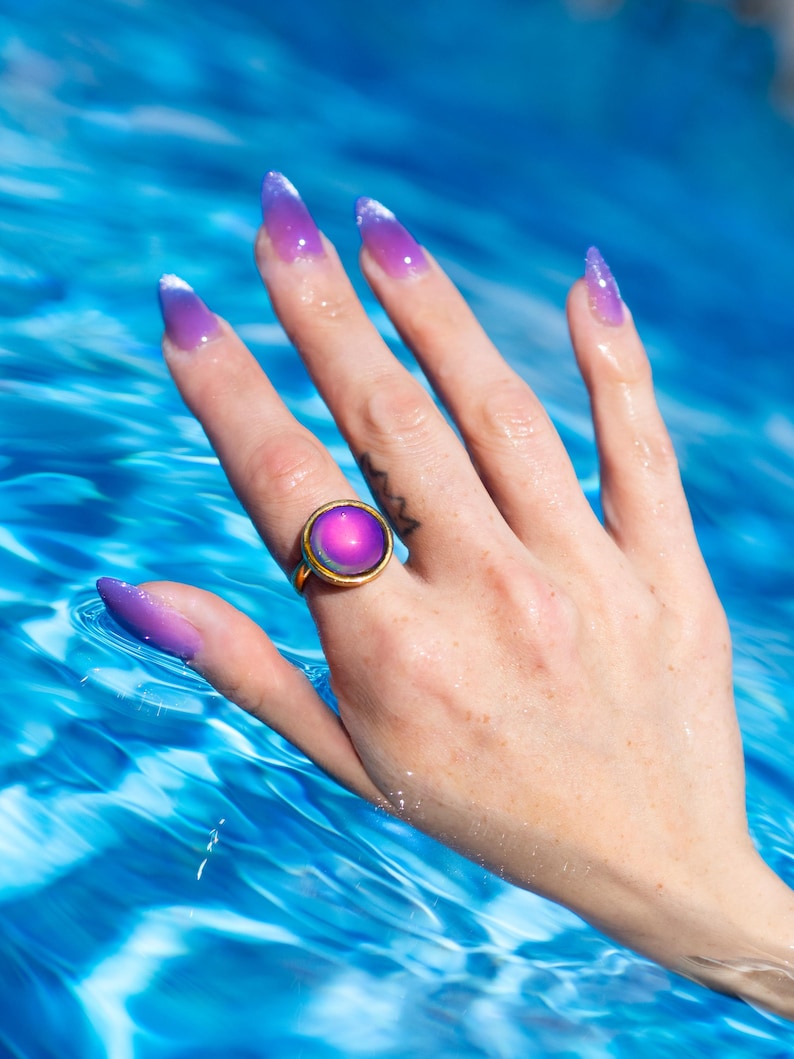 Small gold circle mood ring worn on hand in water from swimming pool, long purple nails on hand
