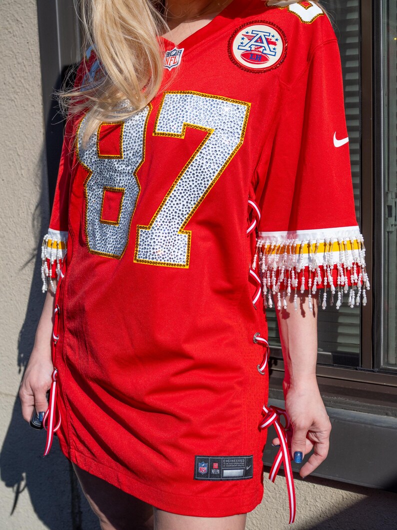 May include: Red football jersey with the number 87 in silver and white. The jersey has white and yellow beaded fringe on the sleeves. Red and white ribbons lace up the sides. The NFL and Nike logos are visible.