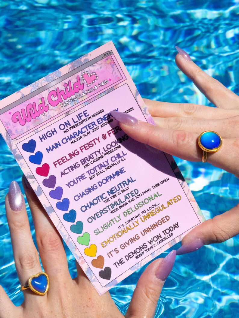 A mood chart card in pink with the text "Wild Child" and a list of moods, floating in a swimming pool with a large silver circle shape mood ring on top with a pink purple stone. The card's text includes phrases like "High on Life" and "Emotionally Unregulated."