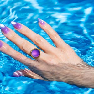 Small gold circle mood ring worn on hand in water from swimming pool, long purple nails on hand