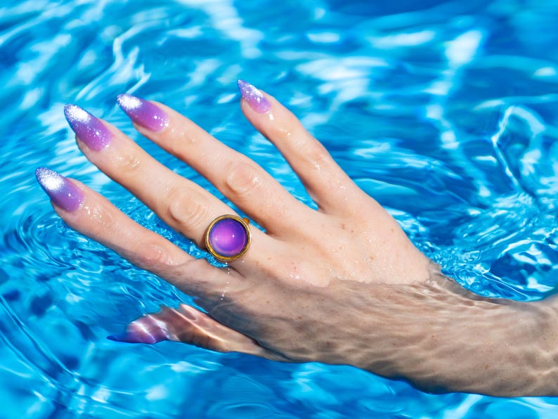Small gold circle mood ring worn on hand in water from swimming pool, long purple nails on hand