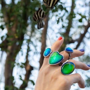 mood rings worn on hand reaching towards a flying butterfly above the hand, rings in silver color with chunky heart and oval shapes in shades of blue and green