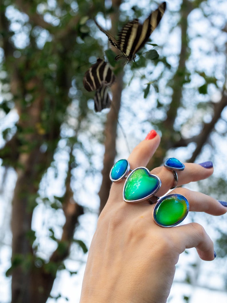 mood rings worn on hand reaching towards a flying butterfly above the hand, rings in silver color with chunky heart and oval shapes in shades of blue and green