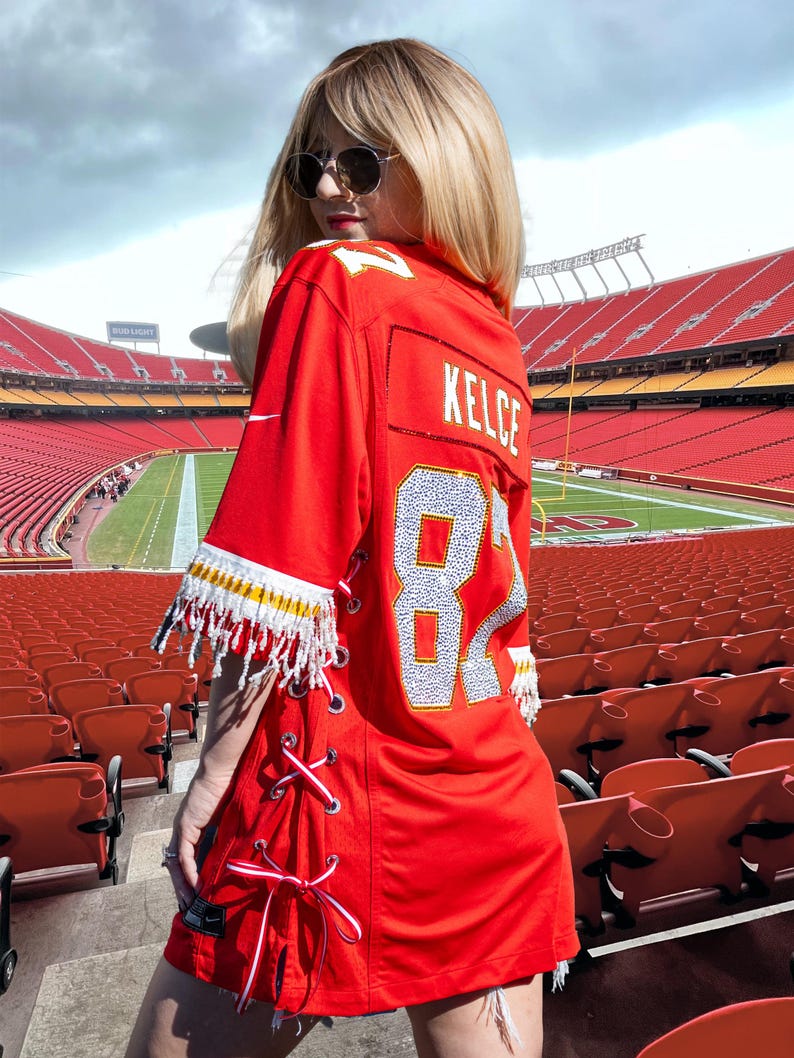 Puede incluir: Vestido rojo de camiseta de f&uacute;tbol con el nombre "KELCE" y el n&uacute;mero "87" en la espalda. El vestido tiene flecos blancos y amarillos en las mangas y un dise&ntilde;o de cordones en los laterales. El vestido se lleva en un estadio.
