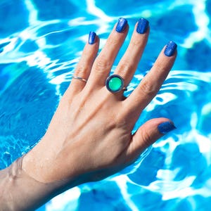 A hand with blue glitter nail polish and a small round silver mood ring with a large green stone. The hand is in blue water, creating a vibrant contrast between the colors displaying the waterproof ability of the rings.