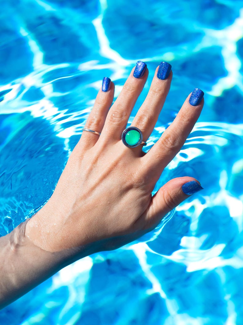 A hand with blue glitter nail polish and a small round silver mood ring with a large green stone. The hand is in blue water, creating a vibrant contrast between the colors displaying the waterproof ability of the rings.