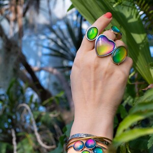 mood rings worn on hand reaching towards leaves, rings in silver color with chunky heart, oval shapes and a gold mood bracelet on the wrist. Mood jewelry is various shades of blue, green, purple, yellow and orange.