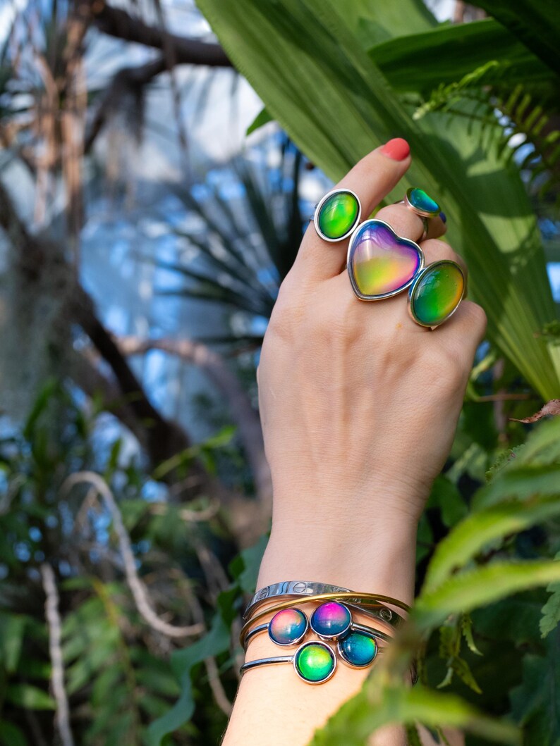 mood rings worn on hand reaching towards leaves, rings in silver color with chunky heart, oval shapes and a gold mood bracelet on the wrist. Mood jewelry is various shades of blue, green, purple, yellow and orange.