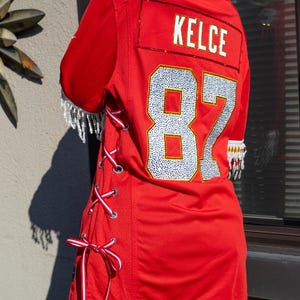 Puede incluir: Una camiseta de f&uacute;tbol roja con el nombre "KELCE" y el n&uacute;mero "87" en pedrer&iacute;a plateada. La camiseta tiene cordones de cinta blanca y roja en el lateral y flecos de cuentas en las mangas.
