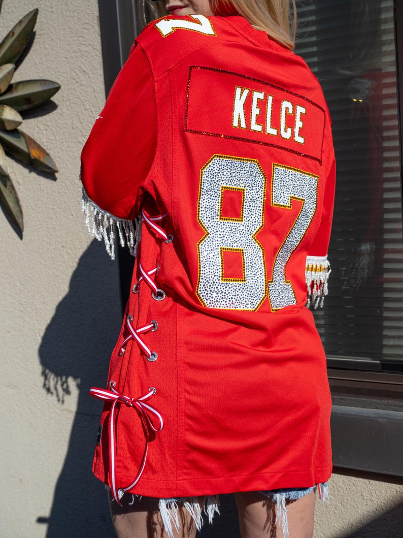 Puede incluir: Una camiseta de f&uacute;tbol roja con el nombre "KELCE" y el n&uacute;mero "87" en pedrer&iacute;a plateada. La camiseta tiene cordones de cinta blanca y roja en el lateral y flecos de cuentas en las mangas.