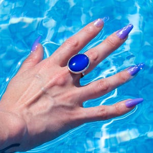 A silver ring with a large, oval, deep blue stone. The ring is on a hand with long, purple fingernails, set against a backdrop of blue water. The water's surface reflects light, creating a shimmering effect.