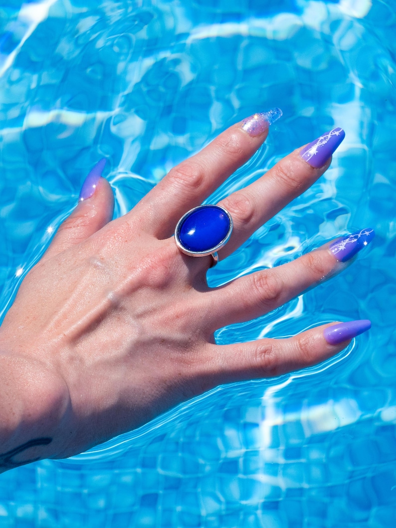 A silver ring with a large, oval, deep blue stone. The ring is on a hand with long, purple fingernails, set against a backdrop of blue water. The water's surface reflects light, creating a shimmering effect.