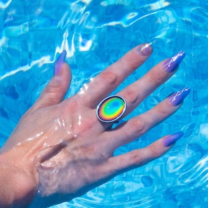 Large silver oval mood ring worn on hand with purple nails underwater in swimming pool, changing color from red on the outside to yellow to green to blue on the inside.