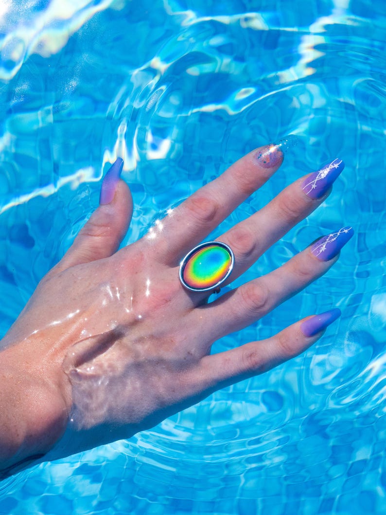 Large silver oval mood ring worn on hand with purple nails underwater in swimming pool, changing color from red on the outside to yellow to green to blue on the inside.