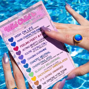 A pink card with the text "Wild Child" and a list of moods, held by a person wearing a gold mood ring with a blue stone. The card's text includes phrases like "High on Life" and "Emotionally Unregulated."