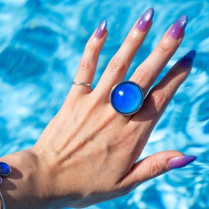 Large silver circle mood ring worn on hand above water from swimming pool, with long purple nails, mood stone is light blue on the edges and dark blue in the center