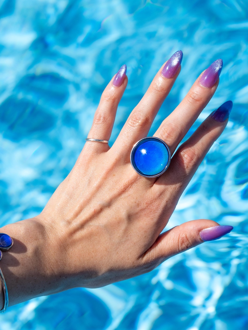 Large silver circle mood ring worn on hand above water from swimming pool, with long purple nails, mood stone is light blue on the edges and dark blue in the center