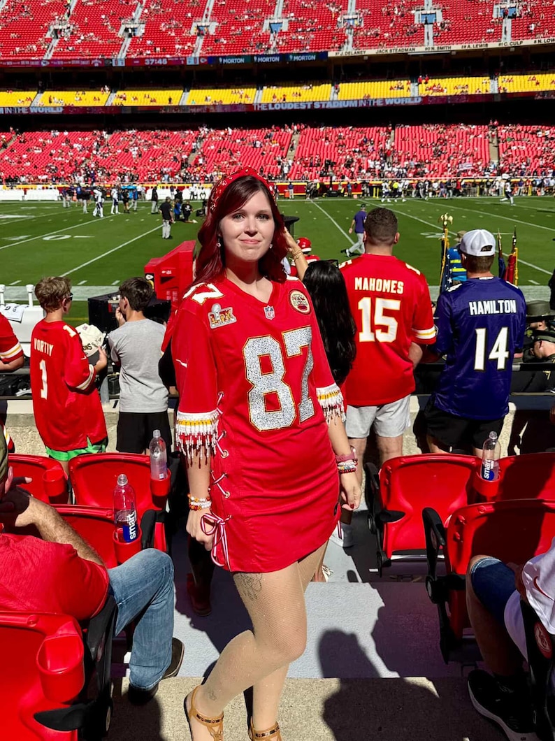 Puede incluir: Un vestido de camiseta de f&uacute;tbol roja con el n&uacute;mero 87 en plata. El vestido tiene flecos blancos y dorados en las mangas. El fondo muestra un estadio de f&uacute;tbol con aficionados y jugadores. El vestido se lleva con sandalias.