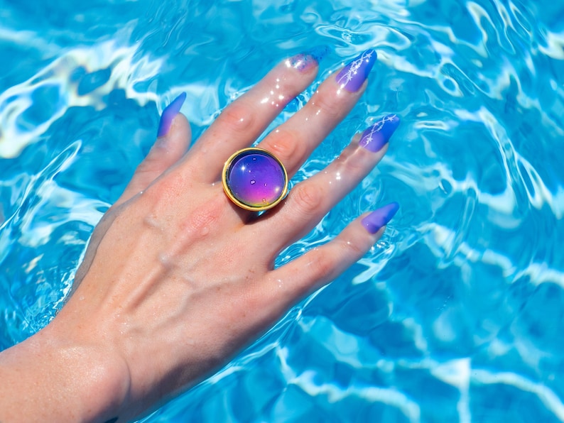 Large gold circle mood ring worn on hand in water from swimming pool, long purple nails on hand